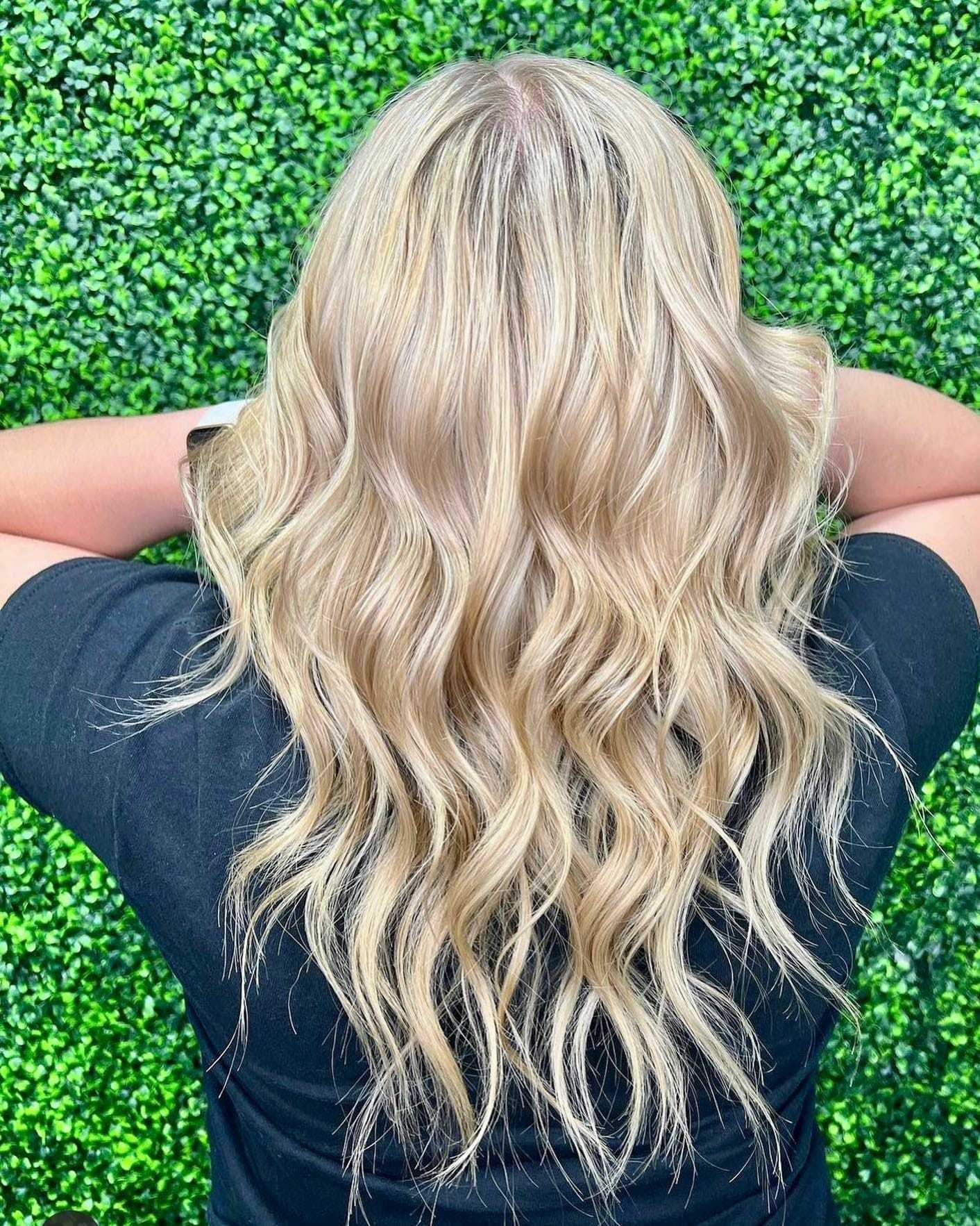 Back view of person with wavy blonde hair against a lush green hedge backdrop.